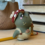 A small handmade clay frog figurine wearing a red mushroom cap and beige overalls, standing on a wooden surface with a yellow pencil and stacked books in the background.