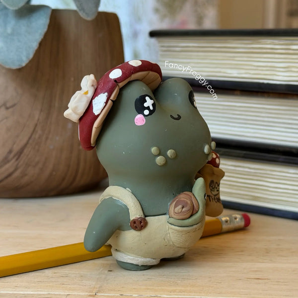 A small handmade clay frog figurine wearing a red mushroom cap and beige overalls, standing on a wooden surface with a yellow pencil and stacked books in the background.