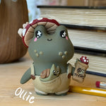 A cute clay figurine of a green frog character wearing a red and white mushroom hat and beige overalls, holding a small bag labeled 'Treats' with a mushroom on it. The figurine is styled on a wooden surface with books and a pencil in the background.