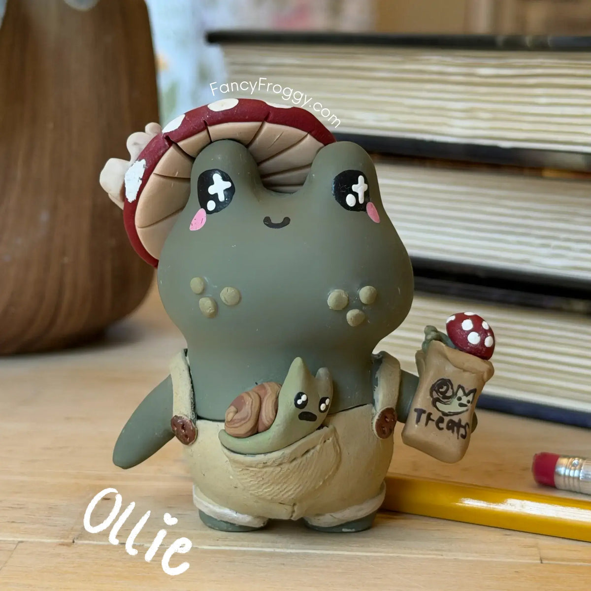 A cute clay figurine of a green frog character wearing a red and white mushroom hat and beige overalls, holding a small bag labeled 'Treats' with a mushroom on it. The figurine is styled on a wooden surface with books and a pencil in the background.
